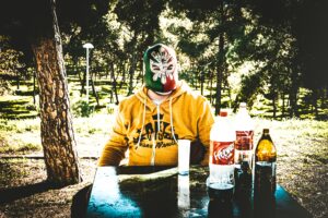 Person in a luchador mask & a hoodie sitting at a table outside with bottles of soda and a 40 in front of them. Person in a luchador mask & a hoodie sitting at a table outside with bottles of soda and a 40 in front of them.